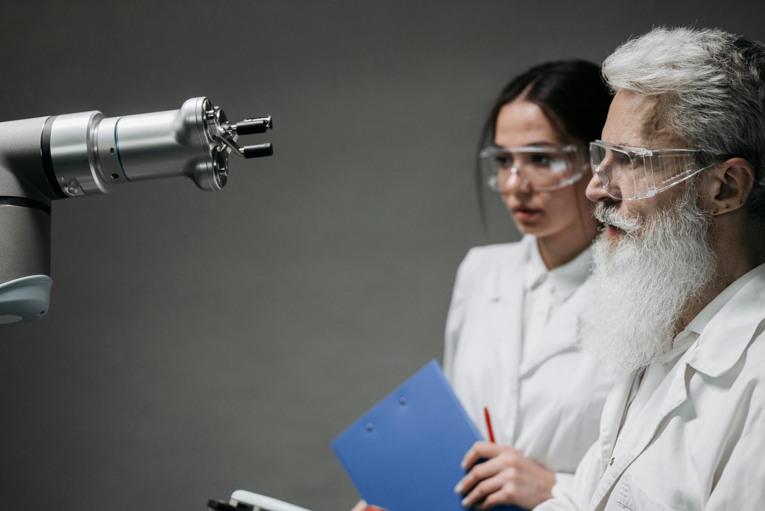 Two scientists wearing lab coats and goggles analyzing a robotic arm in a laboratory setting.