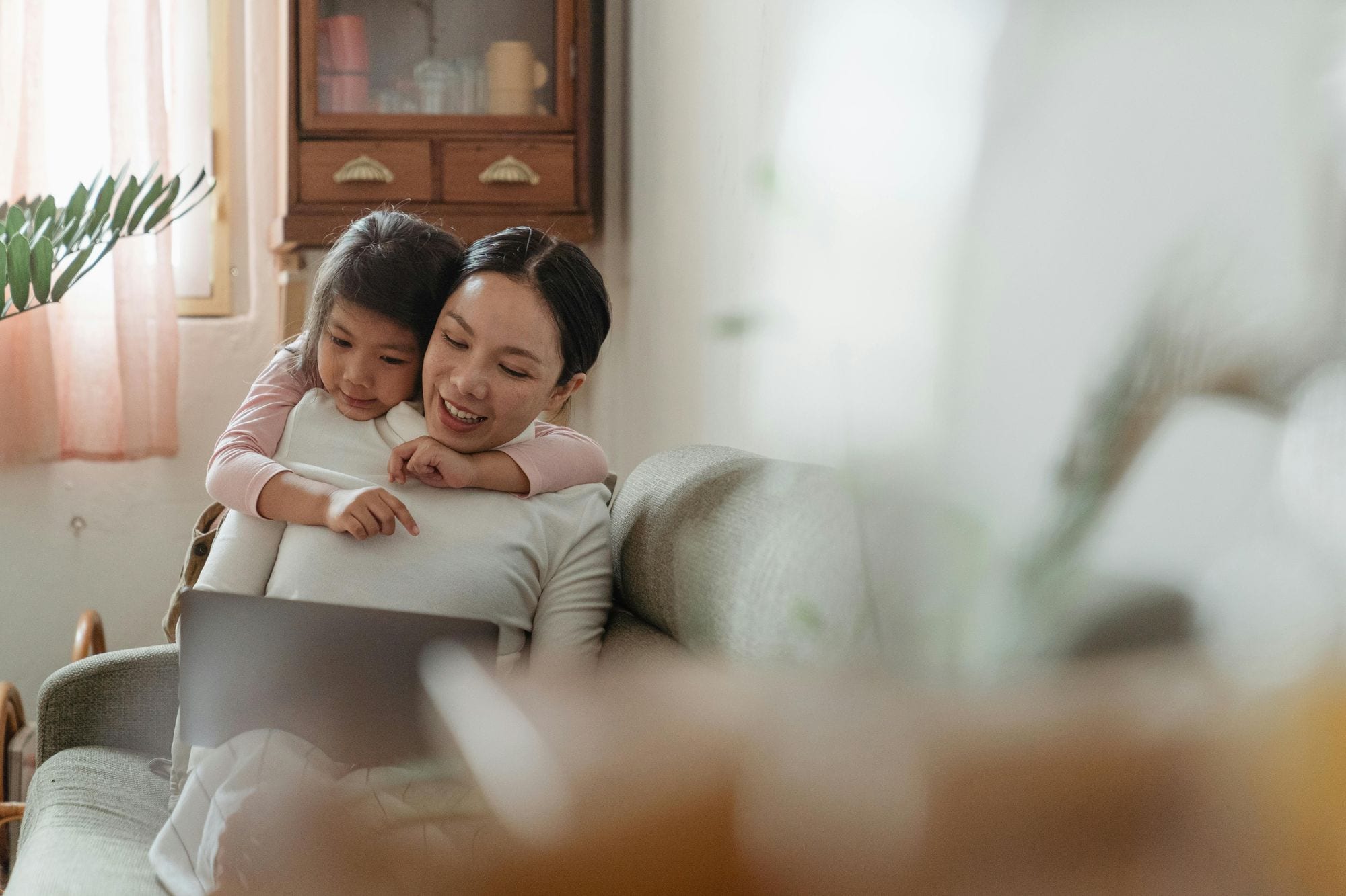Cute little Asian girl in casual clothes embracing cheerful mom surfing modern netbook on cozy sofa in living room